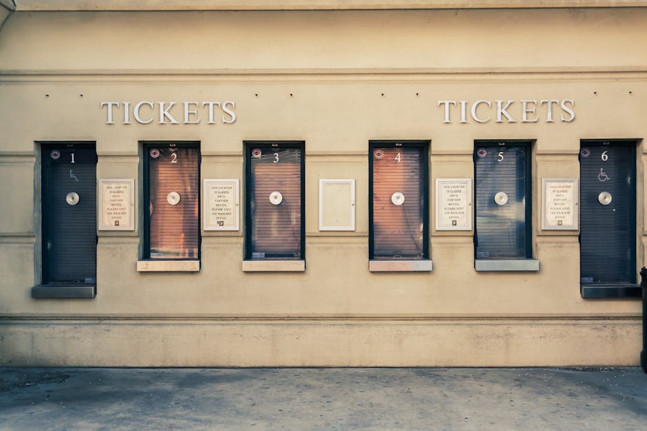 A row of six ticket vending machines mounted on an exterior beige wall, each with a small glass window and a circular payment dial, numbered from one to six. The first, third, and fifth machines display signs with instructions, while the second, fourth, and sixth are covered by dark blinds or shutters. To the left of the machines, there is a white metal plaque with additional information. The ground in front of the machines is paved, and the scene is lit with natural daylight. This image accurately depicts a typical ticket payment area at a public transport station or a parking facility, relevant to house removals or moving services by Man With a Van North Sheen, highlighting the importance of avoiding parking tickets during home relocation or furniture transport activities.