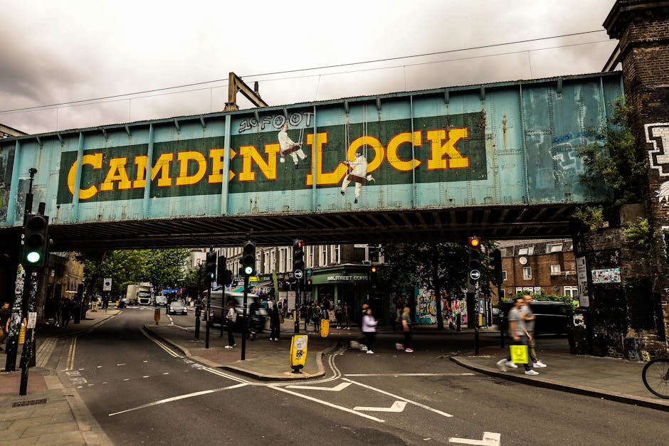 A view of the Camden Lock railway bridge overhead, featuring a large, weathered metal sign with the words 'Camden Lock' painted in bold yellow letters on a turquoise background. The sign is decorated with hanging, painted cutouts of cartoonish people holding onto the letter 'O' in 'LOCK,' adding visual interest. Below the bridge, a busy urban street scene is visible, including traffic lights, cars, and pedestrians crossing at a city intersection. Some pedestrians are walking along the pavement, while others are waiting at the crosswalks. The street is lined with shops and buildings, and the overall scene depicts a typical day in a lively, well-frequented part of North London. The environment is urban with overcast skies, and the overall setting suggests an area that might require local furniture transport or house removals, which Man With a Van North Sheen provides in connection with home relocation and moving services.