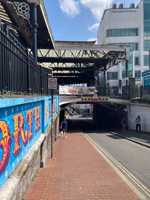 A view of a street scene at Barnes Bridge Station with an underpass beneath a railway bridge. The sidewalk, paved with red bricks, runs alongside a black metal fence adorned with colorful graffiti art. On the left side, there is a blue-painted boundary wall, and above, the railway infrastructure with metal beams and support structures extends over the road. Pedestrians are visible walking along the pavement, some near the underpass entrance. Modern office buildings with glass windows and signage, including one that reads 'International House,' are seen in the background. The sky is partly cloudy, with sunlight illuminating the scene. This setting reflects urban transport and logistics, relevant to home relocation or furniture transport, and may be associated with local removals services such as those provided by Man With a Van North Sheen.