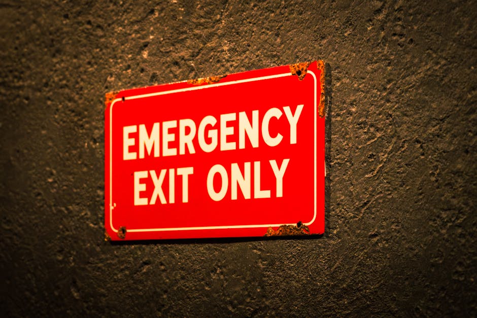 A close-up photograph of a rectangular red metal emergency exit only sign attached to a textured dark wall. The sign features bold white capital letters spelling 'EMERGENCY EXIT ONLY' and has visible rust and chipping along the edges, indicating age and exposure. The sign is positioned slightly off-center to the left, with the wall's rough surface providing a contrasting background. This image likely captures a safety feature commonly found in buildings and properties, possibly near a doorway or exit point, relevant to house removals and moving logistics by Man With a Van North Sheen during a home relocation or furniture transport scenario.