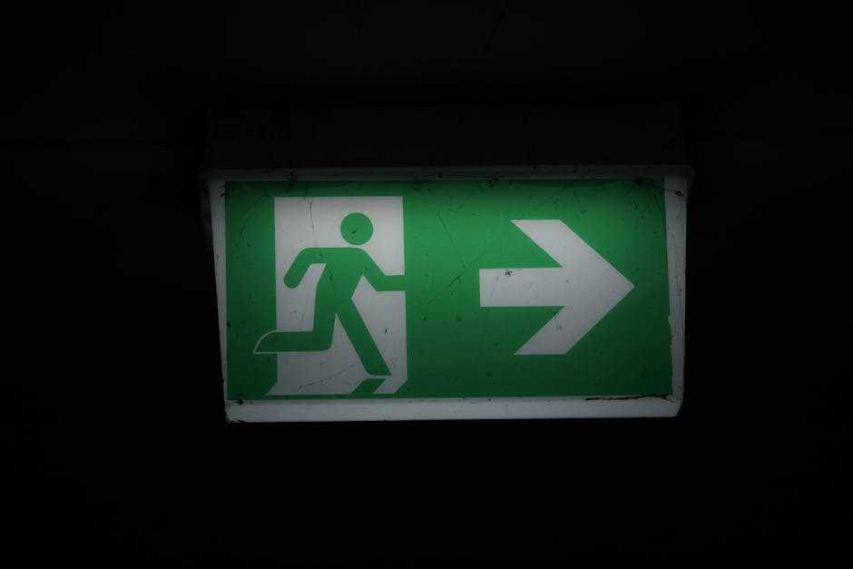 A green illuminated emergency exit sign mounted on a dark wall, featuring a white pictogram of a person running towards a door and an arrow pointing to the right. The sign is positioned inside a building, likely near a corridor or stairwell, indicating the direction for a quick escape route during an emergency. The lighting highlights the sign’s clean design and visibility, which is essential for safe evacuation. This type of signage is commonly seen in buildings involved in home relocation or moving logistics, such as storage areas or building exit pathways, and may be associated with packing and loading processes by services like Man With a Van North Sheen, ensuring safety during furniture transport and home removal activities.