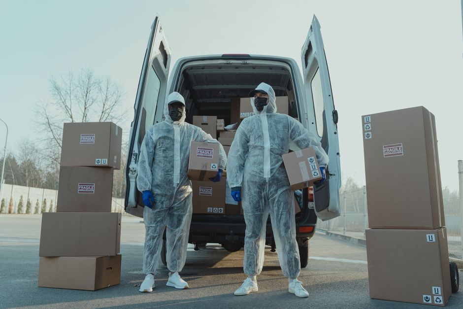 Two movers dressed in protective white coveralls, face masks, and blue gloves stand next to an open delivery van during a home relocation. The van is parked outside on a paved area with a clear sky an
