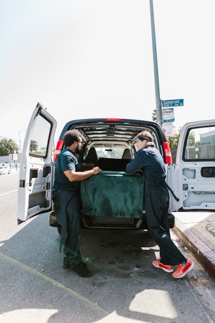 Two movers dressed in protective white coveralls, face masks, and blue gloves stand next to an open delivery van during a home relocation. The van is parked outside on a paved area with a clear sky an