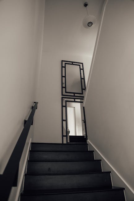 A vertical view of a narrow staircase in a Victorian-style property in North Sheen, showing dark wooden steps and a black metal railing. The staircase walls are painted white, and at the top, there are two geometric black metal frames mounted on the wall, aligning with the stairwell. Natural light is entering from a small window at the base of the staircase, which is visible through an open door at the bottom of the stairs. The ceiling is painted white with a small, simple ceiling light fixture. This scene captures a section of a residential interior during a home relocation process, with no furniture or packing materials visible but highlighting the confined space typical of Victorian flats. Occasionally, ongoing moving operations by Man With a Van North Sheen might involve transporting furniture or boxes through such staircases during packing and loading activities.