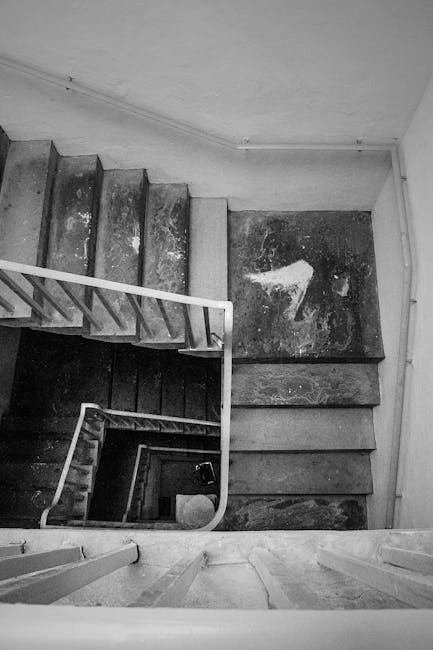 A black-and-white photograph showing a narrow, steep staircase inside a Victorian-style flat in North Sheen, with visible signs of wear and age on the concrete steps and wooden landings, some of which have patches of paint or marks. The staircase is enclosed by plain, smooth walls and features a simple metal handrail on the left side, running along the curved staircase. The image captures an overhead view looking down from the top of the stairs towards the lower floors, highlighting the tight space and the vertical progression typical of older residential buildings. This setting is part of a house relocation scene where careful packing and furniture transport may be involved, as evidenced by the confined corridor and the structural features of the property. Man With a Van North Sheen provides removals and moving services that often handle properties with narrow staircases like this, requiring specialized planning for safe and efficient furniture transport.