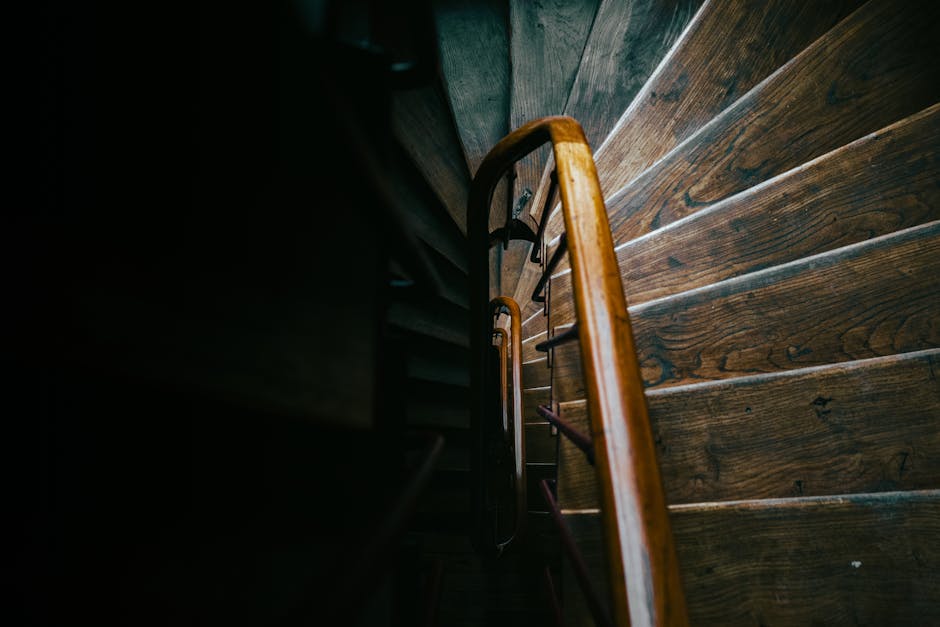 A vertical view of a narrow staircase in a Victorian-style property in North Sheen, showing dark wooden steps and a black metal railing. The staircase walls are painted white, and at the top, there are two geometric black metal frames mounted on the wall, aligning with the stairwell. Natural light is entering from a small window at the base of the staircase, which is visible through an open door at the bottom of the stairs. The ceiling is painted white with a small, simple ceiling light fixture. This scene captures a section of a residential interior during a home relocation process, with no furniture or packing materials visible but highlighting the confined space typical of Victorian flats. Occasionally, ongoing moving operations by Man With a Van North Sheen might involve transporting furniture or boxes through such staircases during packing and loading activities.
