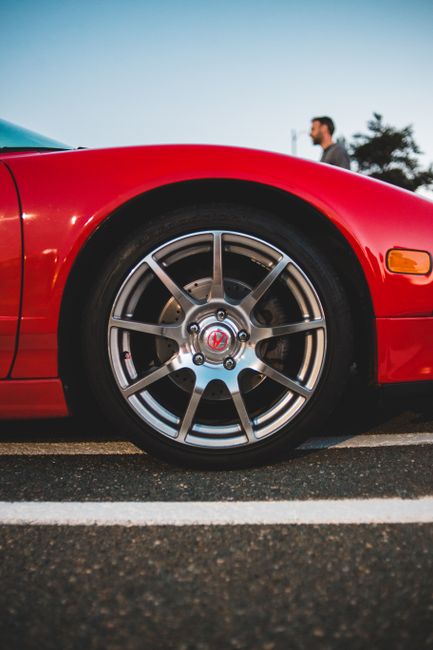 Close-up view of a bright red sports car's front wheel, featuring a multi-spoke alloy rim with a central Volkswagen logo, parked on a paved surface with white parking lines. The vehicle's lower side panel is visible, with a sleek and smooth finish under natural daylight. In the background, slightly blurred, is a man standing outdoors and a tree, suggesting an open area possibly at a relocation or moving site. The scene emphasizes the car's design and condition, relevant to transport and moving services offered by Man With a Van North Sheen, as part of a home relocation process involving vehicle transport or parking considerations.