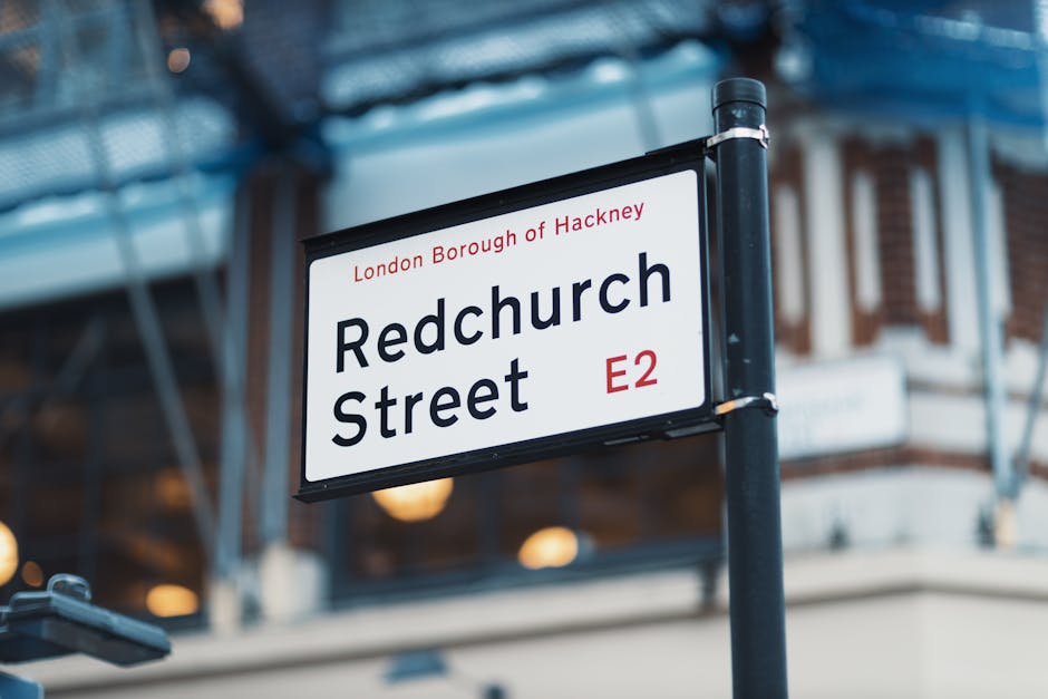 Close-up view of a street sign for Redchurch Street in the London Borough of Hackney, displaying the street name in bold black letters on a white background with red postcode '.E2' in the lower right corner, mounted on a black metal pole. In the background, parts of a building with brick and white facades, along with a glass canopy or roof structure, are visible, illuminated by warm ambient lighting. The image captures the sign at street level during daylight, with the focus on the sign indicating its relevance for residential and commercial moving logistics related to house removals and furniture transport in the area, highlighting an urban environment suitable for professional relocation services such as those offered by Man With a Van North Sheen.