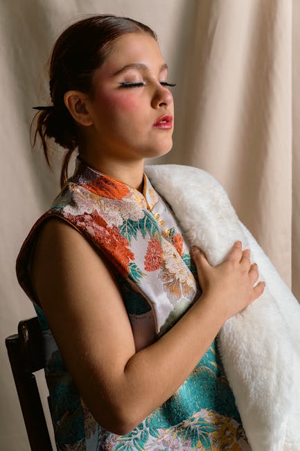 The image shows a young woman with her eyes closed and a peaceful expression, sitting indoors near a cream-colored curtain. She has dark brown hair pulled back and is dressed in a colorful floral kimono-style top with shades of orange, teal, and beige. She is holding a large, white, fluffy pillow or blanket close to her chest with her right arm, while her left hand rests gently on it. The lighting is soft and natural, highlighting her serene facial expression. The background is neutral, with the curtain providing a calm and understated backdrop. The scene conveys a sense of comfort and relaxation, which can be associated with home environment and personal space. This image could be relevant for content related to home comfort, relaxation, or personal well-being, complementing topics about house removals or moving services offered by Man With a Van North Sheen, as it emphasizes a peaceful state before or after a home relocation process.