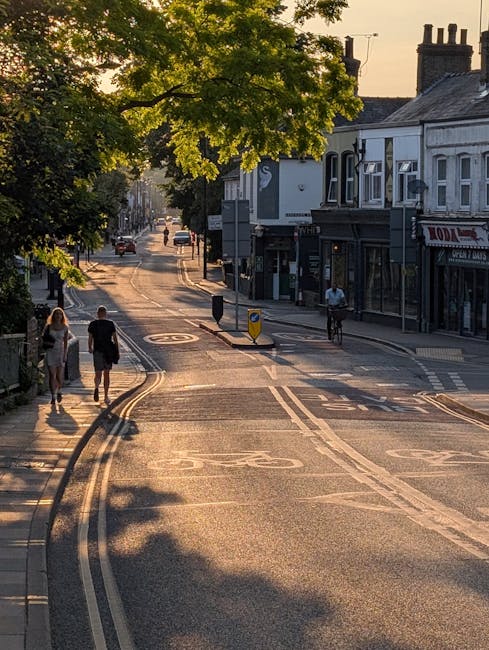 A quiet street scene in North Sheen during late afternoon or early evening, with a large green tree partially shading the road. The street is lined with small shops and buildings, including a white storefront with large windows and a dark-colored upper floor. Two pedestrians, a man and a woman, are walking along the sidewalk on the left, while a cyclist rides on the right side of the street. Several cars are visible in the distance, and road markings such as double yellow lines, traffic islands, and bus lane indicators are present on the tarmac. Shadows cast by the tree and buildings create patterns on the road, and the overall scene emphasizes calm urban residential and commercial activity, relevant to house removals or moving services in North Sheen, as managed by Man With a Van North Sheen.