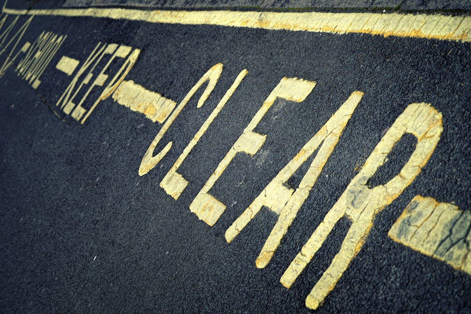 A close-up photograph of a black asphalt road surface with yellow painted capital letters spelling 'CLEAR' and part of another word. The letters are slightly worn and cracked, indicating frequent use or weathering. The image features a double yellow line running parallel above the painted text, and the scene appears to be taken outdoors during daylight, with natural lighting highlighting the texture of the asphalt and the paint. In the background, there is a section of a curb or pavement, with a slight blur, suggesting the photo focuses on the road markings. This visual is relevant to house removals and moving services, as it depicts road signage that might be encountered during furniture transport or loading processes, which [COMPANY_NAME], such as Man With a Van North Sheen, considers when planning home relocation routes.