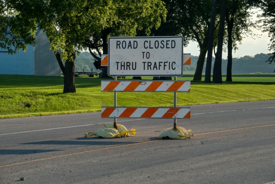 A rectangular road closure sign with black text reading 'ROAD CLOSED TO THRU TRAFFIC' is mounted on a metal frame reinforced with orange and white diagonal striped barriers. The sign is positioned on a paved street, with two sandbags at its base to secure it in place. Behind the sign, there are lush green grass patches and a row of tall trees providing shade. In the background, a large light blue industrial building is visible, along with a trailer and distant hills under a partly cloudy sky. The scene suggests a temporary road closure possibly related to infrastructure work, with the barriers indicating restricted access, which may impact house removals or relocation logistics managed by [COMPANY_NAME], such as street closures and skip license arrangements for local moves in North Sheen.