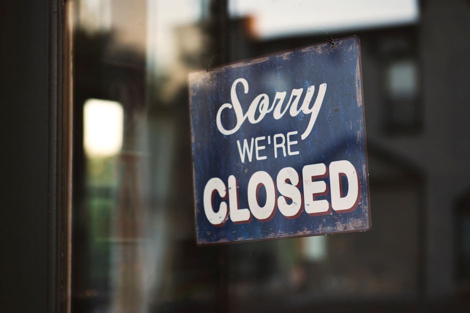 A close-up photograph of a blue metal sign with white cursive and bold block lettering that reads 'Sorry We're Closed.' The sign appears weathered, with visible chipping paint and rust around the edges. It is mounted on a glass door or window, reflecting a blurred background of a residential street with nearby buildings and soft natural daylight. The image captures a quiet moment indicating the closure of a business or service establishment, possibly related to house removals or relocation services provided by Man With a Van North Sheen, situated in North Sheen, London. The focus is on the sign, emphasizing its message with a subtle implication of service downtime, relevant to the context of moving and home relocation activities that might involve temporary closures or planning of moving logistics.