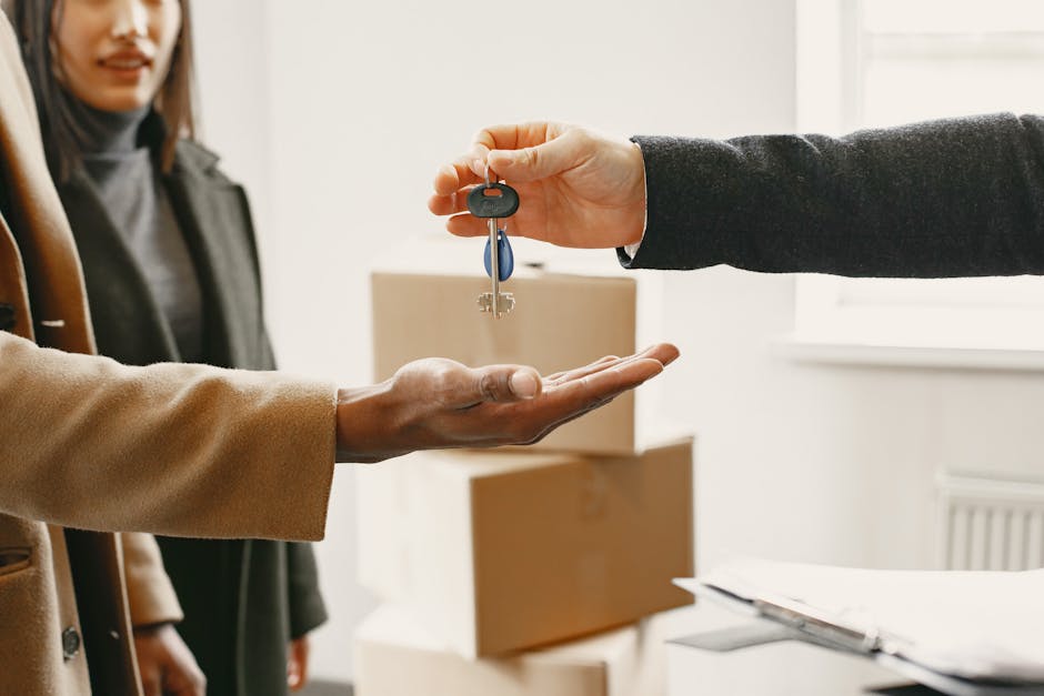 A person in a dark suit is handing over a set of keys to another individual with a tan coat inside a room during a house move, with a woman observing in the background. The room features a window allowing natural light, and a stack of cardboard boxes and packing materials are visible on a table in the foreground. The scene depicts a formal handover related to property occupancy, with the keys symbolising access, and it is set during a home relocation process overseen by Man With a Van North Sheen, which offers removals and packing services. The background includes a radiator and a light-colored wall, emphasizing the indoor environment. This visual relates to the final stages of a house move, such as tenant handover or property transfer, highlighting key exchange procedures typical in residential removals and property management.