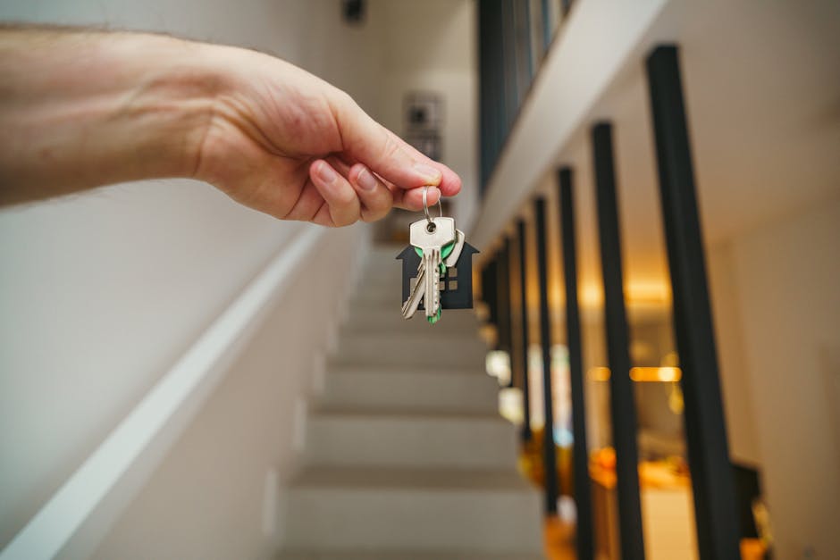 A close-up of a person's hand holding a set of keys with a small black plastic keychain, positioned in front of a staircase inside a residential property. The staircase has light-colored steps and a black metal balustrade with vertical bars, leading upwards. The interior is well-lit with warm ambient lighting that reflects off the wooden handrail and the walls painted in neutral tones. In the background, part of a hallway or living space is visible, indicating an interior setting typical of a home undergoing a property handover or fresh occupation. The image captures the moment of key transfer or house identification, relevant to house removals, packing, and home relocation processes, as exemplified by Man With a Van North Sheen’s services for tenant handover repairs after a move.