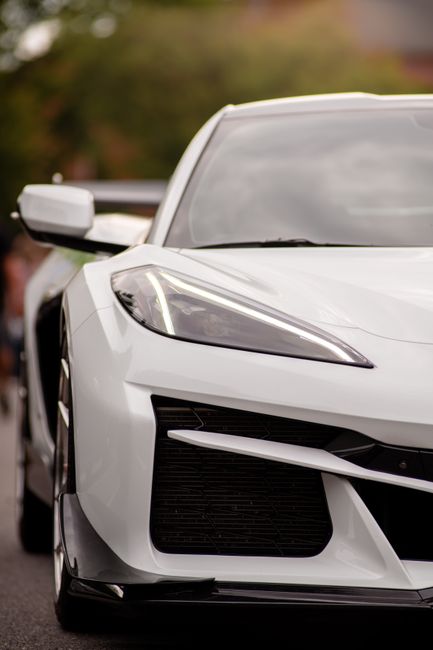 A close-up view of the front left side of a white modern car, showing the angular headlight, black grille, and side mirror. The vehicle is parked on a pavement with a blurred background of trees and b